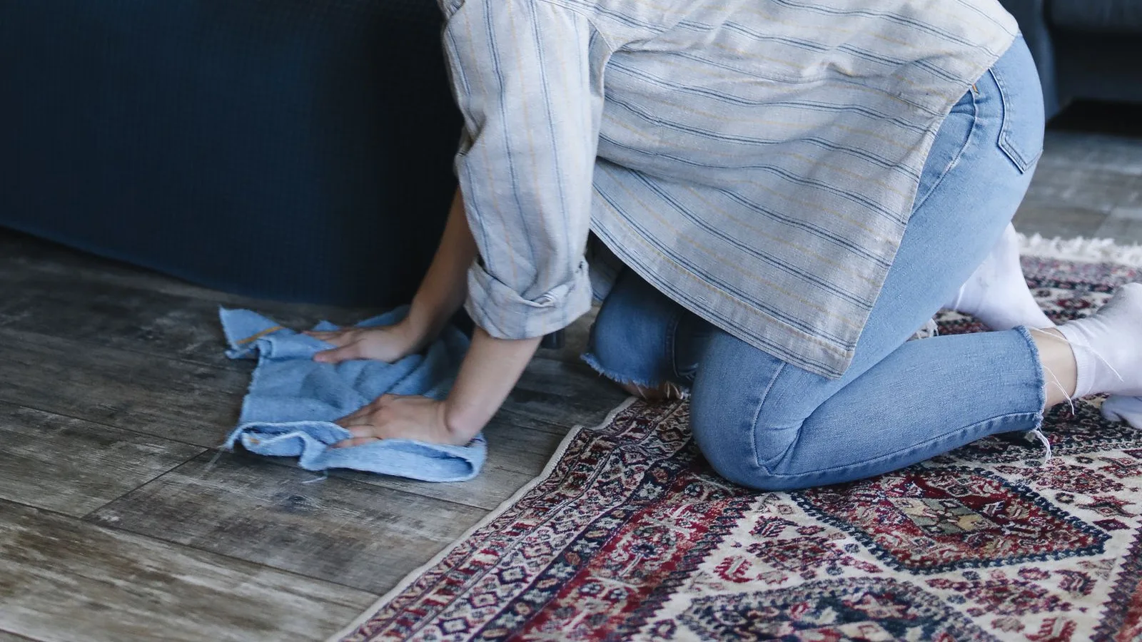 A person kneeling on a patterned rug, pressing a damp cloth onto the floor while spot cleaning by hand