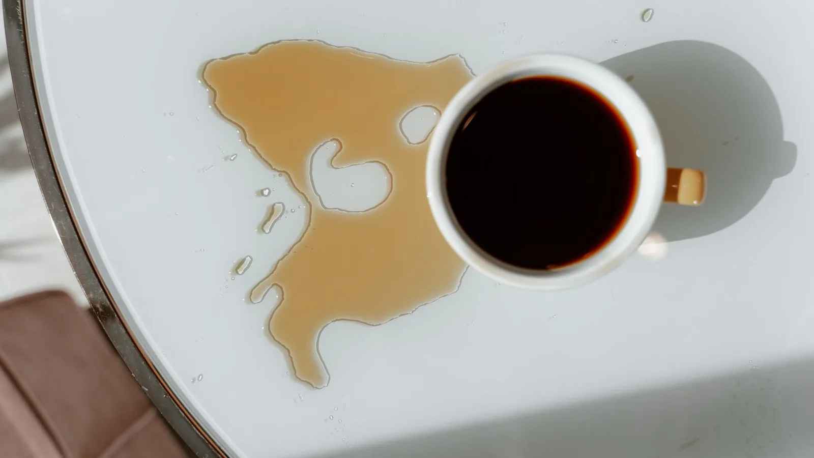 A spilled cup of black coffee pooling on a white table in warm morning light
